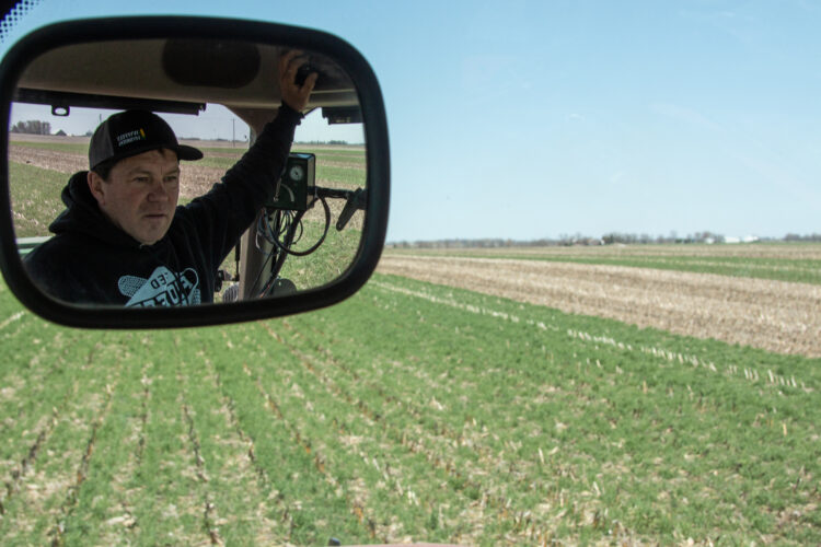 Farmer Brian Hora seen through rearview mirror in the cab of a seed planter as he seeds a field green with cover crops.