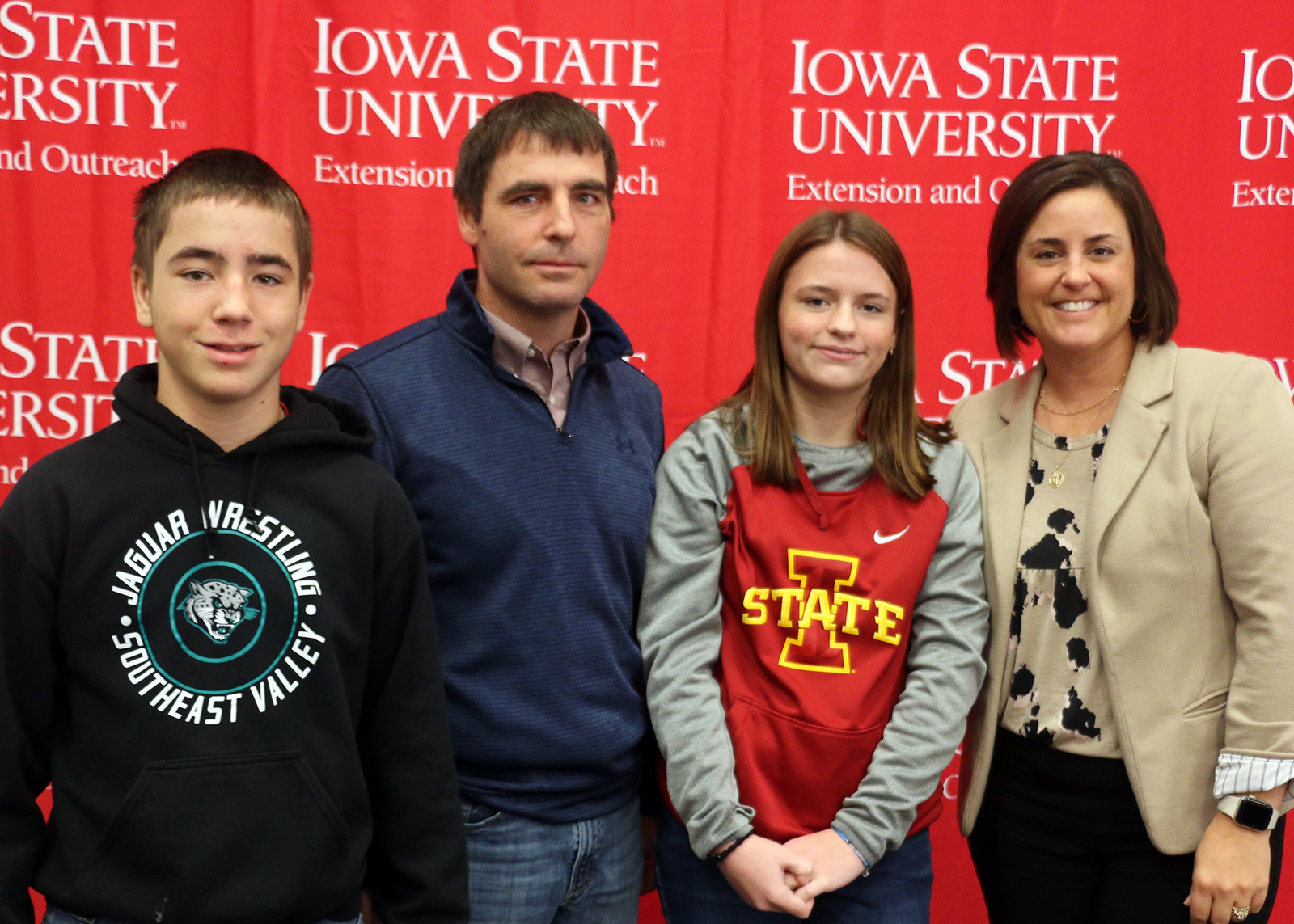 Kellie Blair smiles with her two children and husband in front of an Iowa State University banner back-drop.