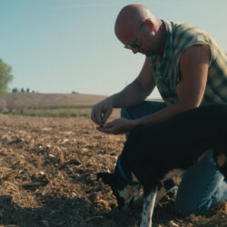 Iowa regenerative farmer Kelly Garrett kneels in a crop field, looking at a handful of soil.