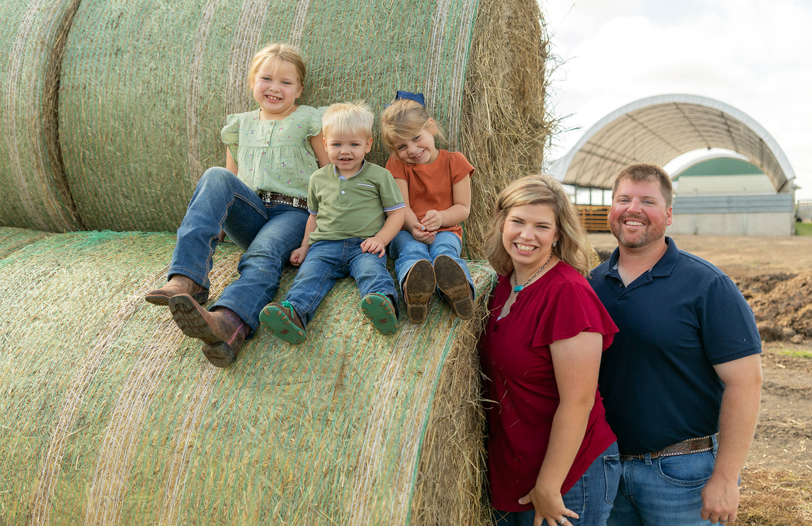 Sara Preston smiles with her husband and three children in an outdoor family photo on their farm. The children sit atop a hay bale. Sara and her husband stand next to them.